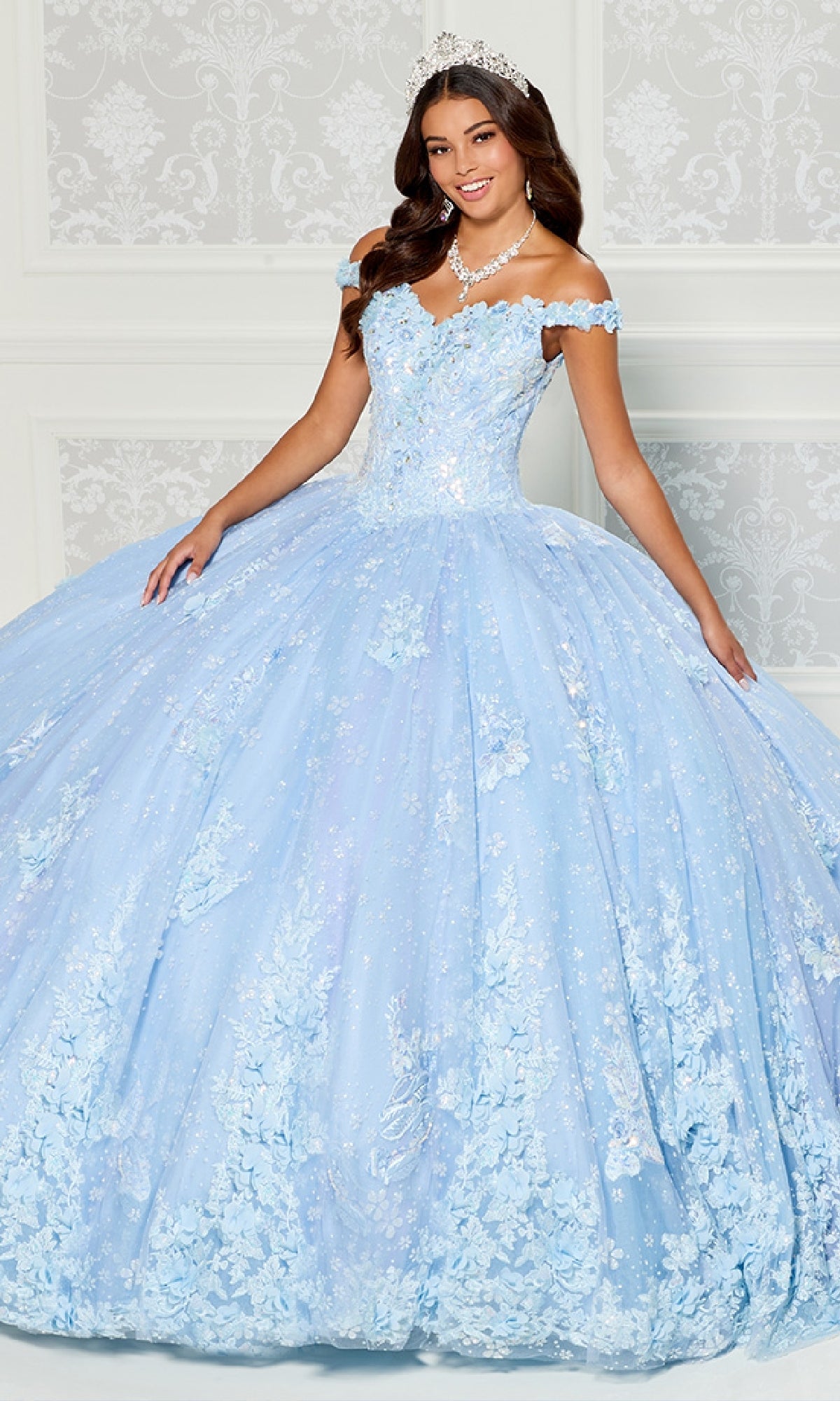 A young woman stands against a pale patterned background wearing the Quinceanera Dress PR30111 By Princesa, a light blue off-the-shoulder gown with floral lace embroidery, complemented by a tiara and jewelry.